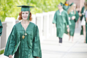Young woman in green commencement attire, walking towards camera with diploma