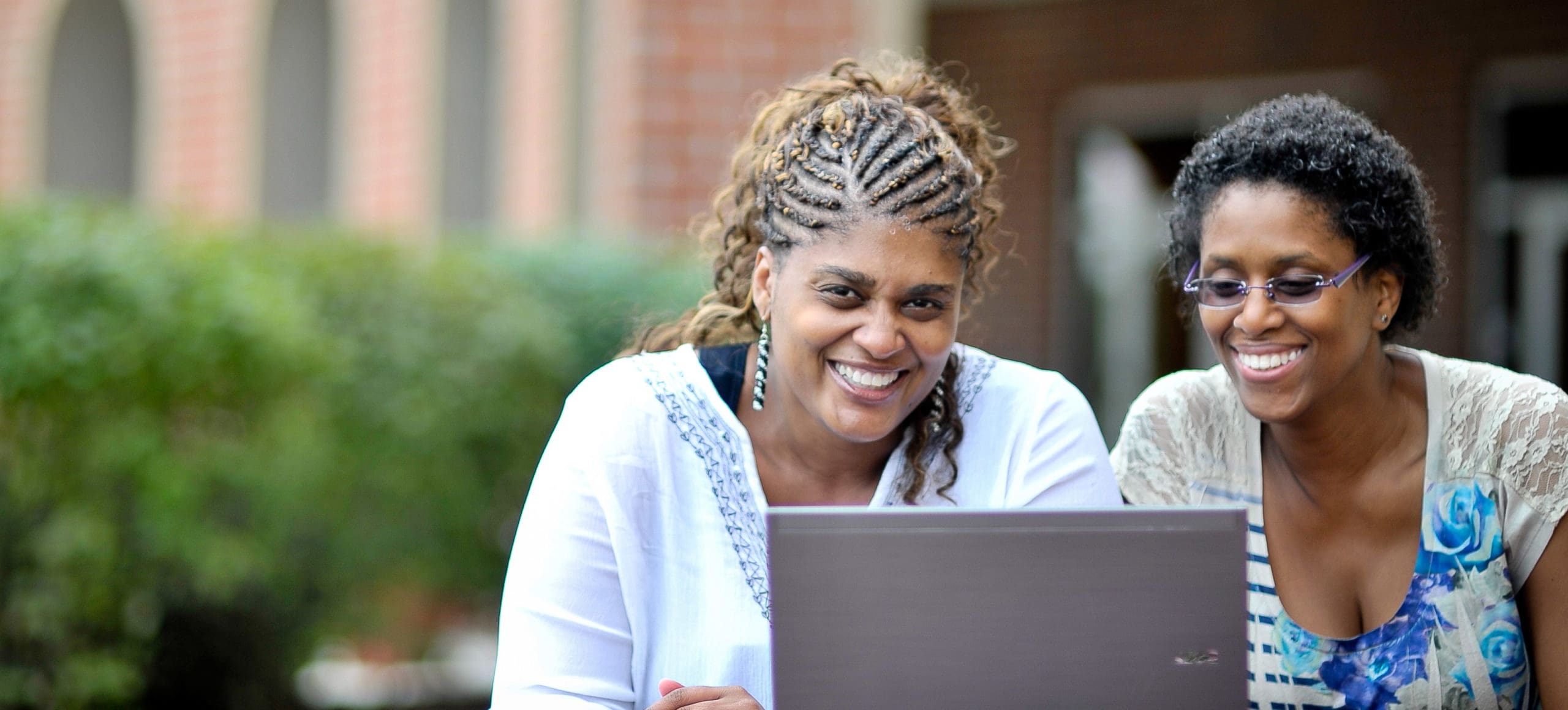 AU_Hero_Images_2x_GSLC_Veterans › Antioch University 2 smiling women on laptop outside GSLC campus