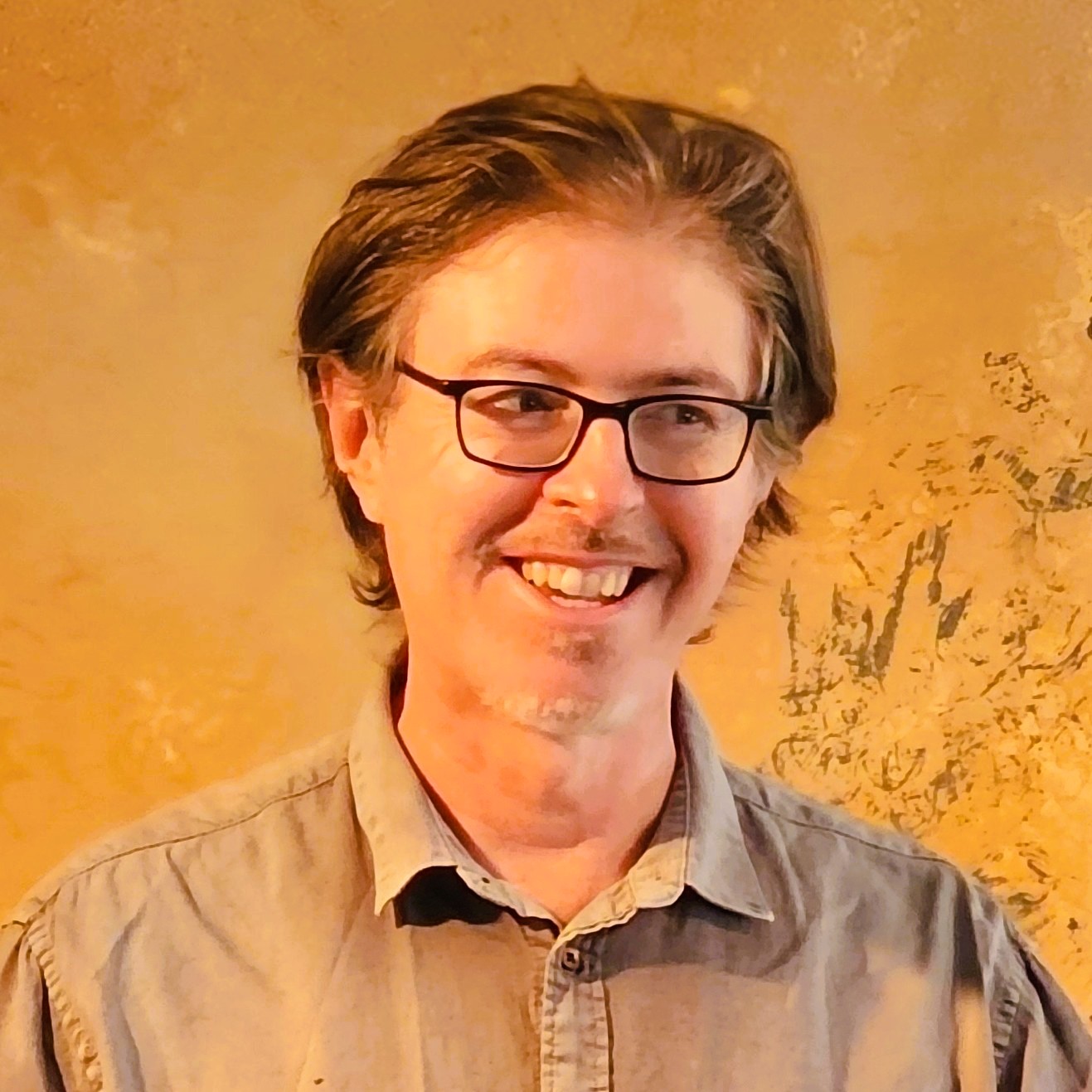 Alistair McCartney 2026 | Antioch University Alistair McCartney, orange background, guy smiling with glasses and blonde hair
