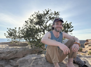 image | Antioch University Smiling man in a sleeveless shirt and cap sits on rocky ground outdoors, with trees and blue sky in the background.