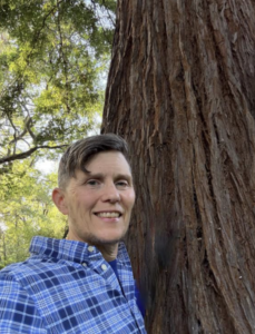 image | Antioch University Person in a blue plaid shirt smiles at the camera while standing beside a large tree trunk in a park.