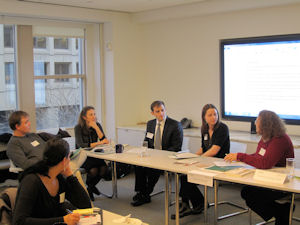 Six professionals seated around a conference table in a meeting room, discussing as a slide is projected on a wall screen behind them.