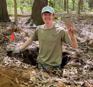 image | Antioch University Man in a green shirt and cap excavating in a leaf-covered forest, smiling and showing a peace sign at the camera.