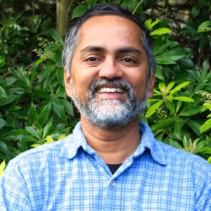 Gopal Krishnamurthy, smiling in a blue button up shirt with greenery behind him.