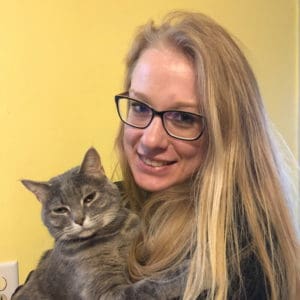 Melody Rossiter smiling and holding their cat against a yellow background