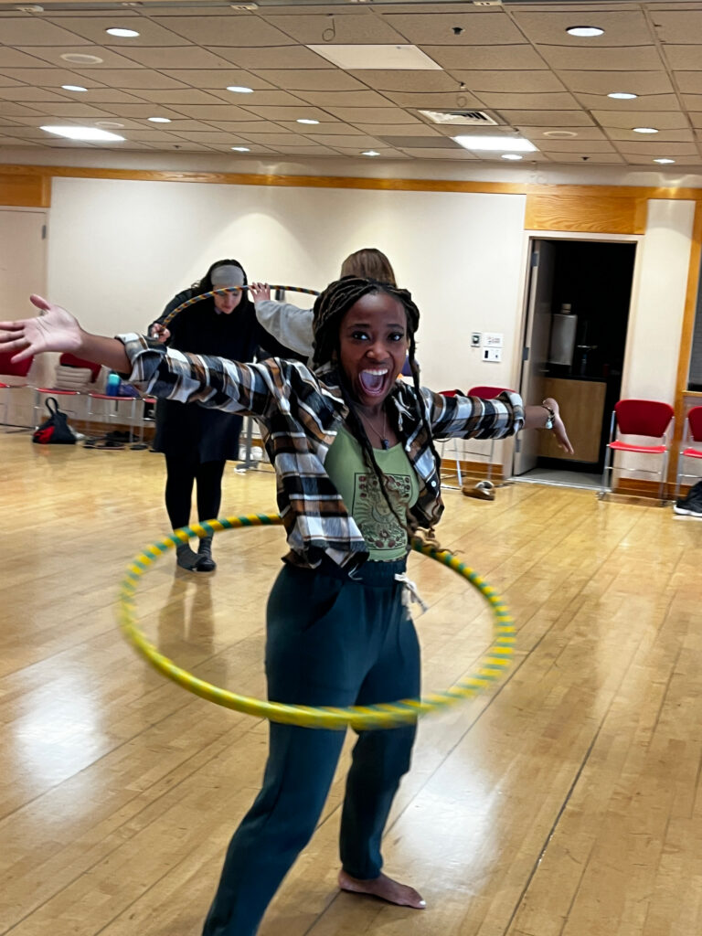 A person enthusiastically hula hooping with arms outstretched in a large indoor room, smiling toward the camera.