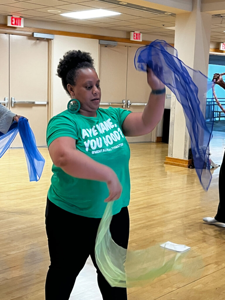 A person in a green shirt moving colorful sheer scarves with their hands in a dance or movement activity inside a studio.