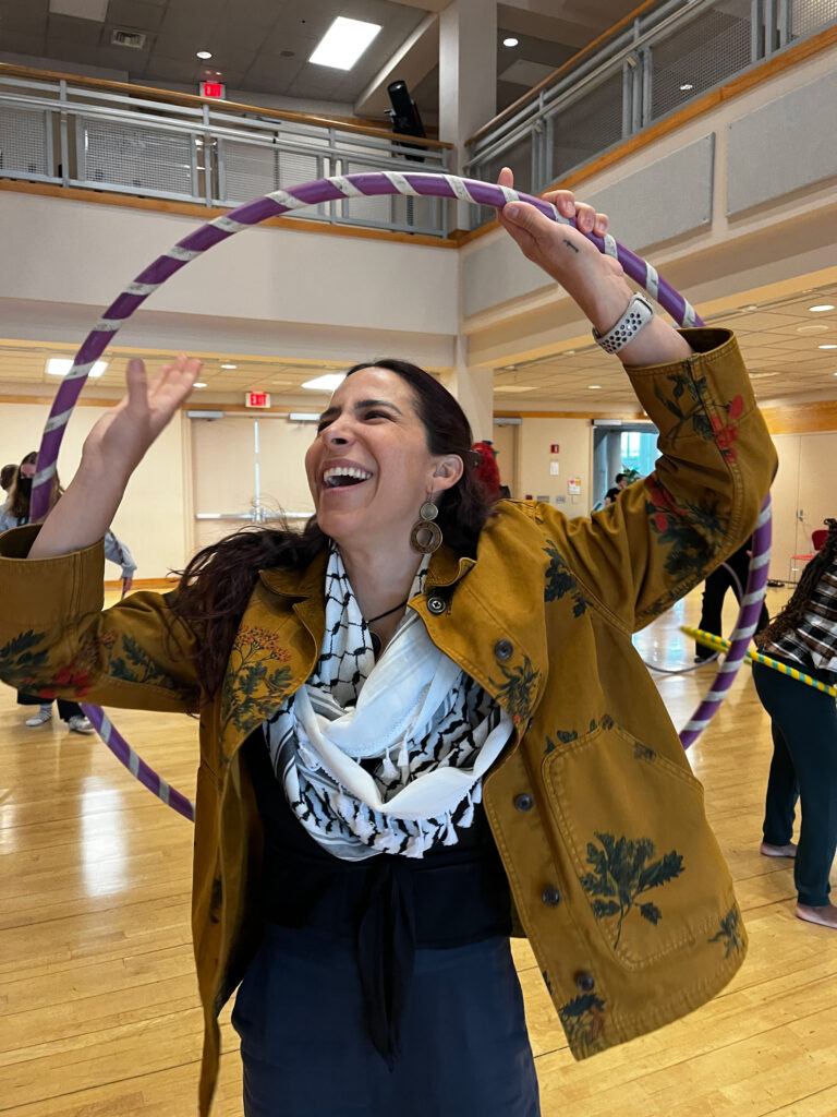 A woman laughing while holding a purple hula hoop above her head in a bright indoor studio space.