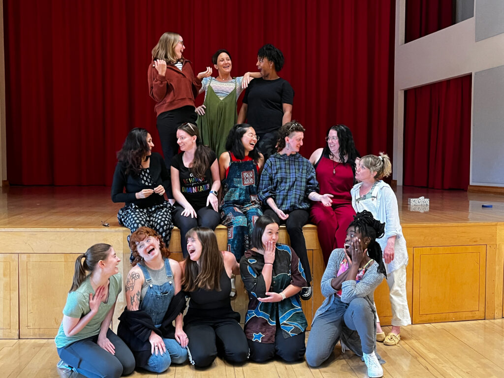 A group of adults sitting and kneeling together on a stage in front of a red curtain, smiling, laughing, and interacting playfully with one another.