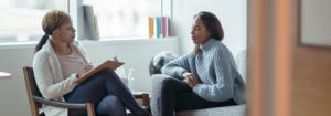 A Therapist meets with her female client in her office. The client is seated on a sofa with her arms across her body as she looks visibly nervous. The Therapist is seated in a chair in front of her as she talks about what to expect from the appointment and takes notes on her clipboard.