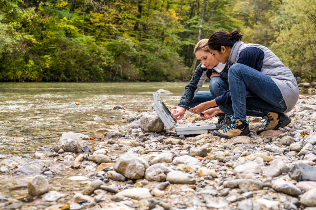 Low angle view of female biological researchers squatting at riverside and discussing measured data. Analyzing water quality in mountain river. Environmental protection, biological research concept.