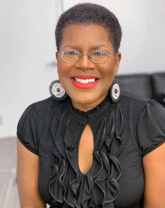 TananariveDue Antioch University Smiling woman with short natural hair glasses red lipstick wearing a black ruffled blouse and large circular earrings