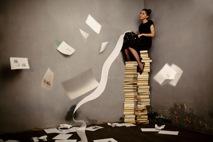 Woman sitting on top a tall stack of books typing.