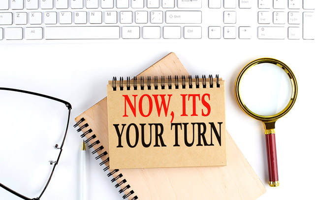 Notebook that says, Now its your turn on a table with glasses and a magnifying glass.