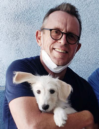 Charley Lang- Man in blue shirt holding a white puppy in from of a blue wall.