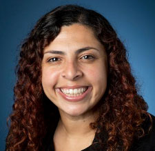 Esther Gobrial, MS, LMFT- This image is a close-up portrait of a smiling woman. She has brown layered hair that falls in ringlets around her face, and she is wearing clothing with black and brown colors.