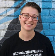 Manny Kemphues- Person with short hair wearing a black shirt with white letters reading, #SchoolsNotPrisons, in front of a white and blue brick wall