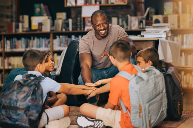 Person sitting in a chair with 4 kids wearing backpacks. They all have their hands together in the center of the circle.