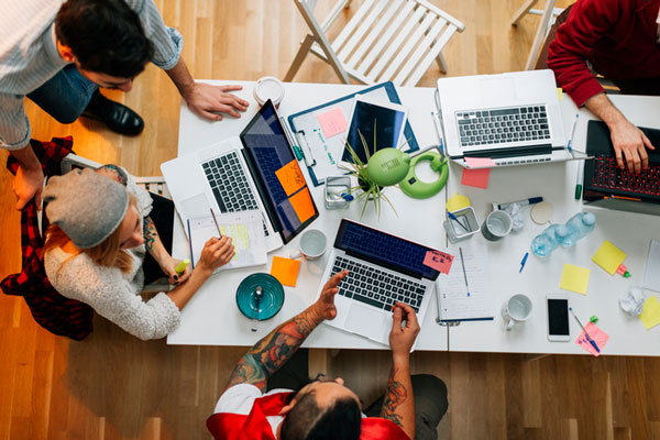 An overhead view of people having a writing workshop on a white desk. There are 4 people, 4 laptops, and various office supplies.