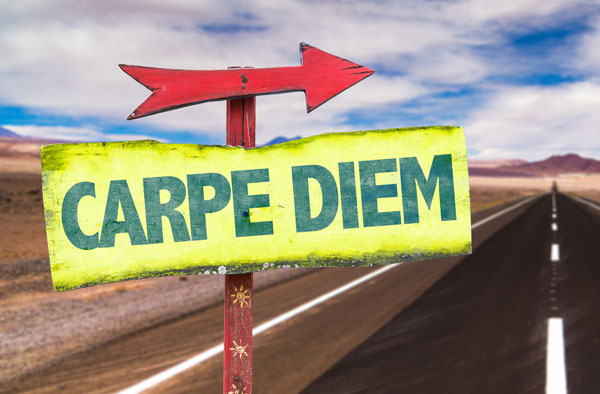 Road sign with a red arrow above a yellow sign that says, Carpe Diem in green. In the background there is highway, desert, and a blue sky with clouds.