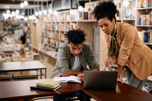 Teacher in beige blazer and scarf helping student in library on computer