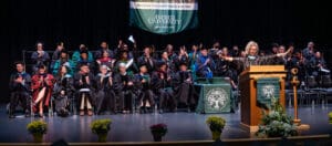 laura-andrews-director-of-instituitional-advancement › Antioch University Stage with graduates and a speaker. Graduates are wearing a variety of regalia. There are green signs that say Antioch University and the speaker is at a podium.