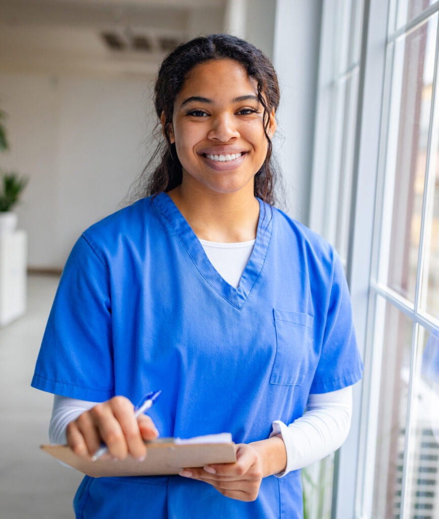 Nursing student in blue scrubs holding a chart and smiling.