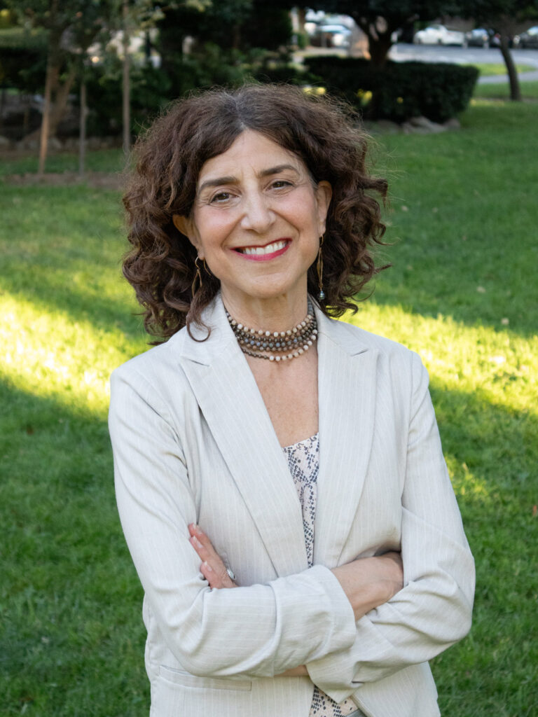 LV-Headshot-2025 › Antioch University A woman in a white blazer standing in front of out of focus grass with dappled light behind