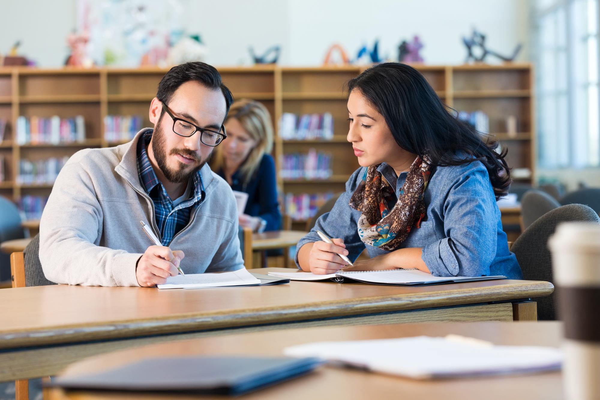 Two students studying together at a library desk, writing in notebooks and discussing their work, with bookshelves behind them.