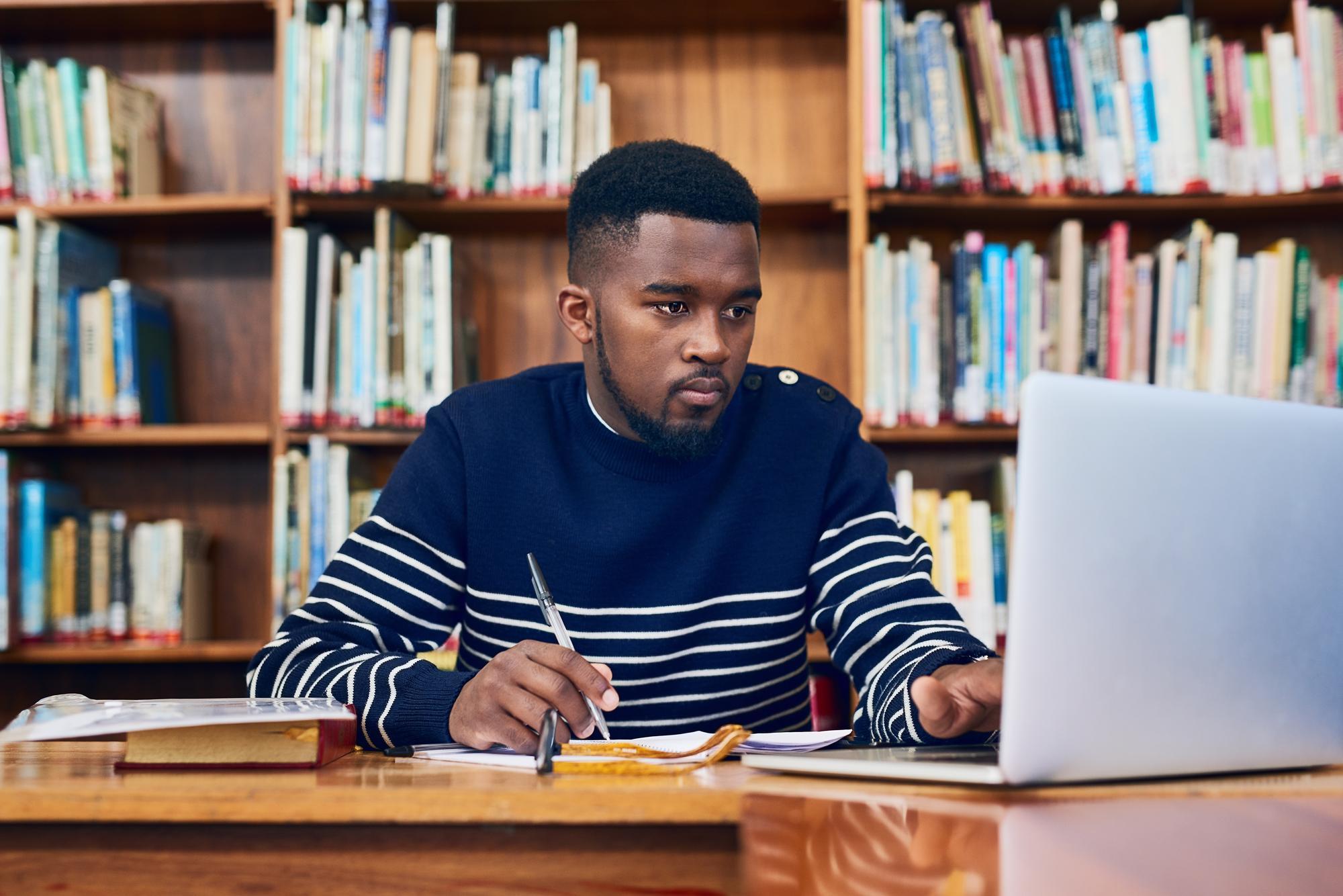 Student studying at a library table, working on a laptop while taking notes, with bookshelves in the background.