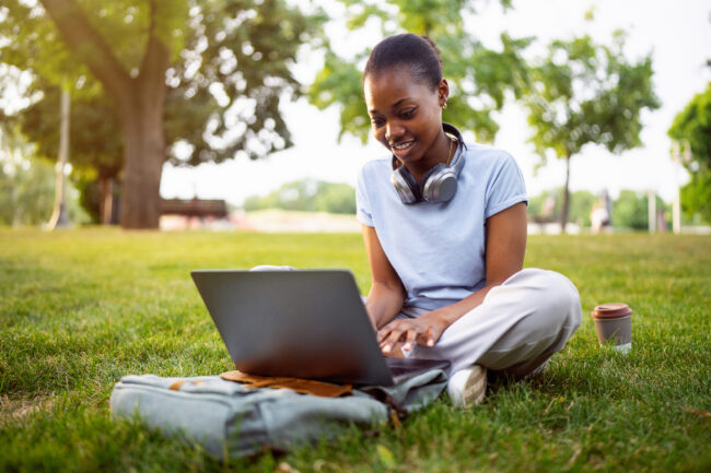 Woman sitting cross‑legged on park grass, typing on a laptop with headphones around her neck and a backpack beside her.