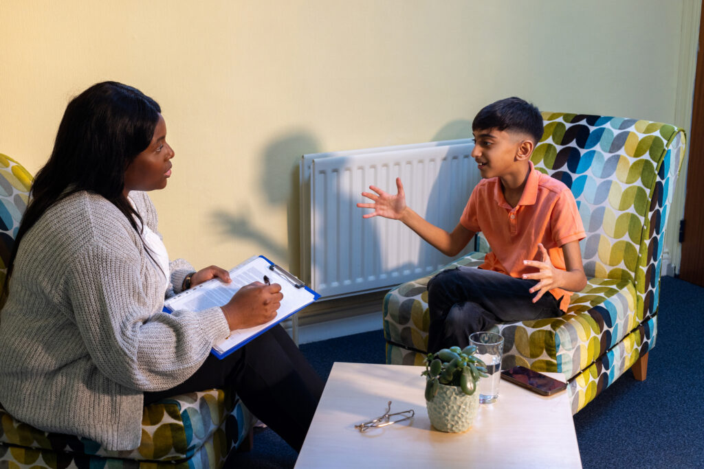 Focused Counselling Session with Young Boy | Antioch University Woman with clipboard speaking to a boy seated on a colorful patterned chair in a cozy room.