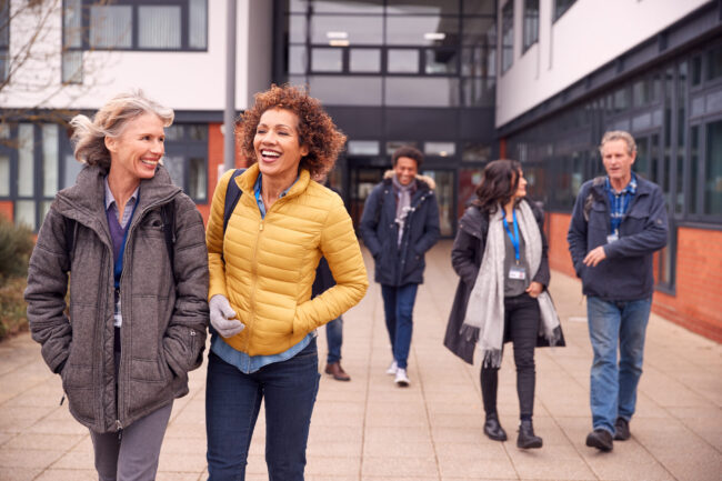 Group of diverse students walking outside a campus building, smiling and chatting; two women in the foreground, one in a yellow jacket.