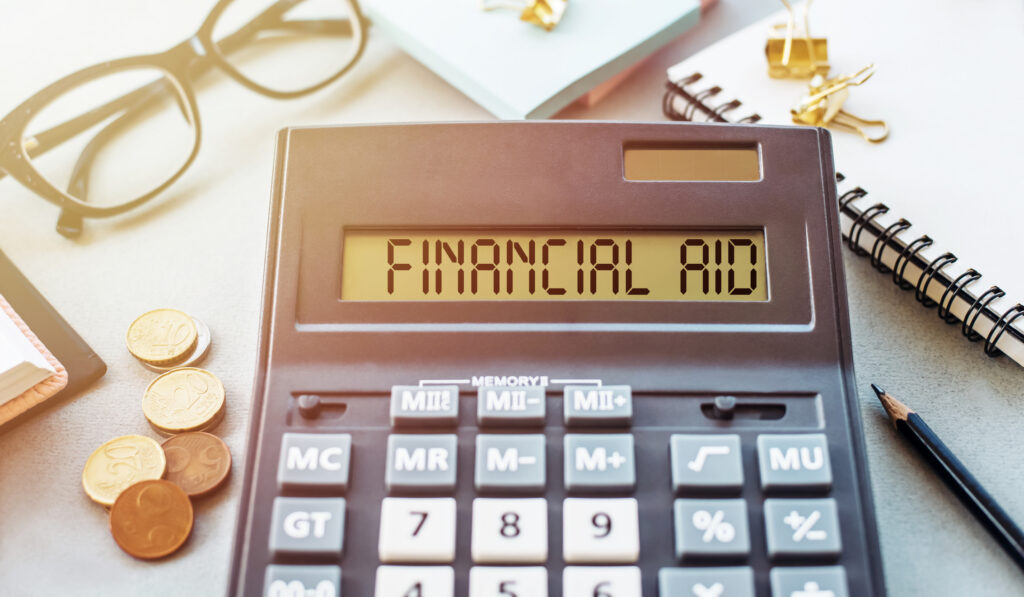 Calculator showing 'FINANCIAL AID' on the display, surrounded by glasses, coins, a notebook, and a pencil—implying financial planning or aid calculations.