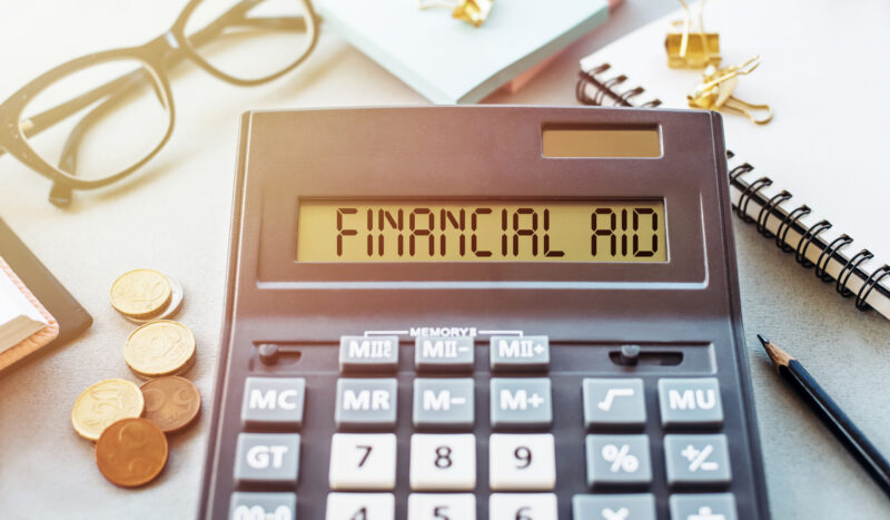 Calculator showing 'FINANCIAL AID' on the display, surrounded by glasses, coins, a notebook, and a pencil—implying financial planning or aid calculations.