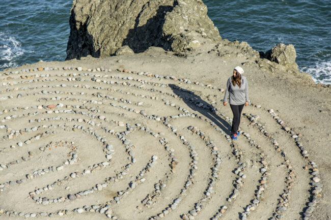 Woman walking along a circular stone labyrinth drawn in the sand by the ocean.