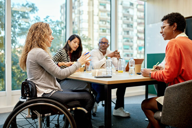 Four colleagues in a meeting around a table; a woman in a wheelchair speaks while others listen and take notes with water bottles and laptops nearby.