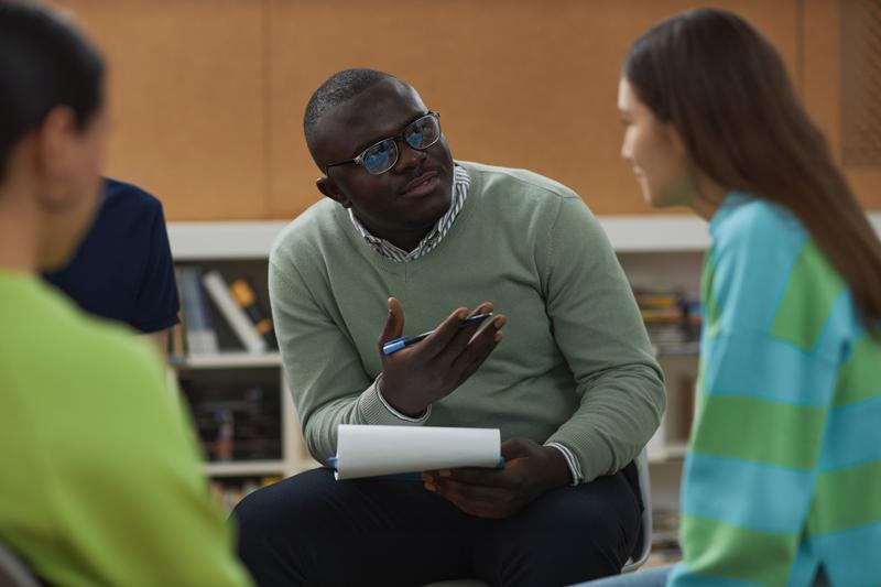 Group study discussion: a man in glasses holds a pen and notebook while talking with two peers in a library setting.