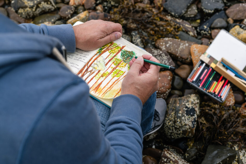 Drawing in a Sketch Book | Antioch University Person sketching a colorful forest scene in a notebook on a rocky beach, with colored pencils nearby