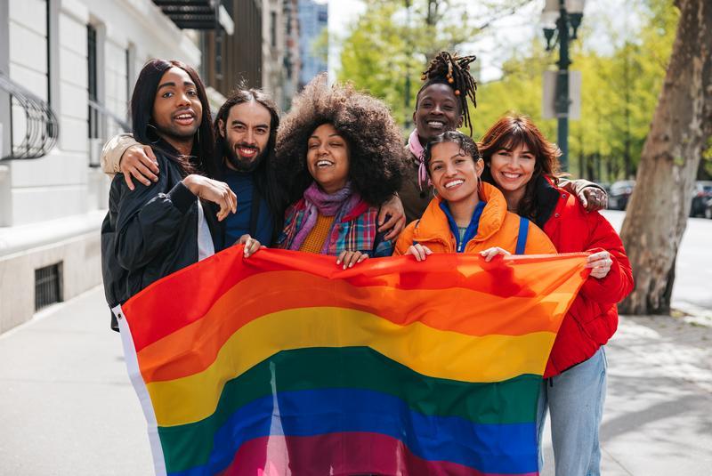 Group of friends posing with a rainbow flag before the LGBTQIA+ Pride Parade | Antioch University Six diverse friends smile and pose with a large rainbow Pride flag outdoors in a city street.