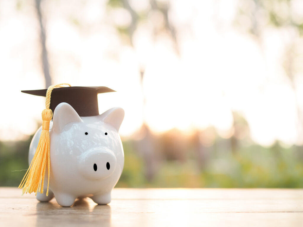 White piggy bank wearing a black graduation cap with a yellow tassel, outdoors on a wooden surface