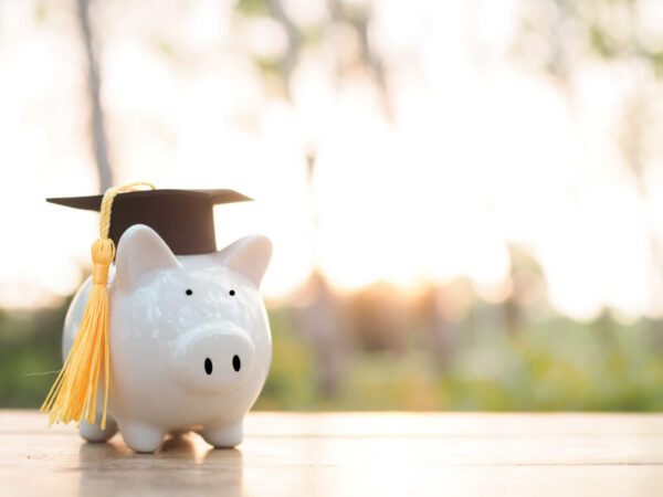 White piggy bank wearing a black graduation cap with a yellow tassel, outdoors on a wooden surface