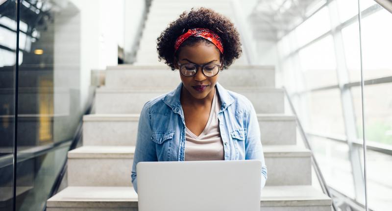 Young woman with glasses and a red headband sits on indoor stairs using a white laptop, wearing a light beige top and a denim jacket in a bright modern setting.