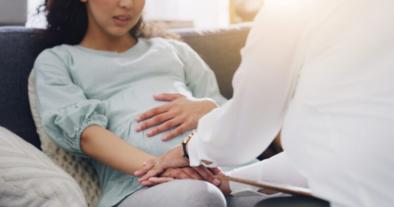 Pregnant woman relaxing on a couch while a healthcare professional holds her hands for support and care.