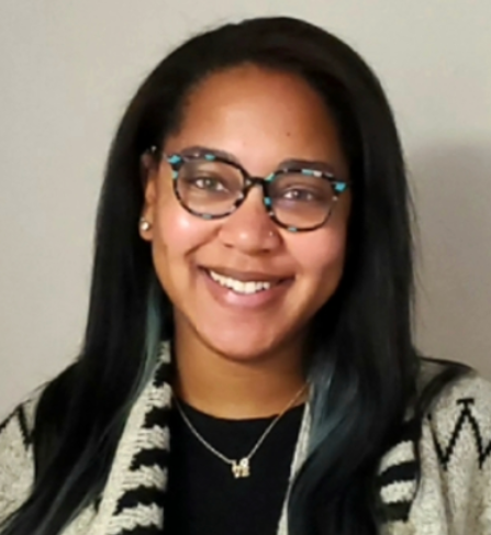 Dr Camille Black | Antioch University Smiling woman, long dark hair, blue glasses, silver necklace, striped coat, black shirt