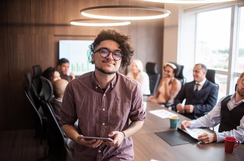 Young man with glasses smiles and holds a tablet at the head of a conference table in a modern office. Behind him, coworkers discuss and work on charts and documents at a long meeting table.