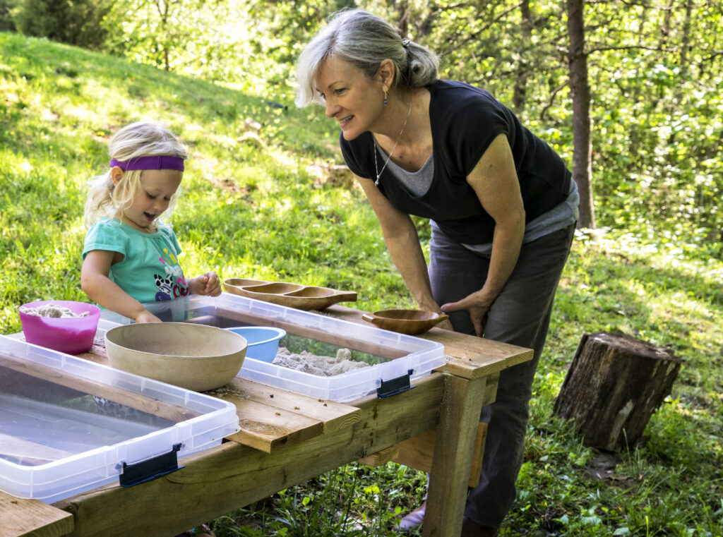 Adult caregiver leaning over a child as they play with sand in clear bins on a wooden table outdoors in a green garden.