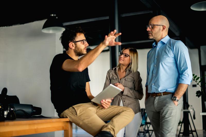 Three colleagues discuss a project: a man in a black shirt with a tablet explains, while a woman and another man listen in a modern office.