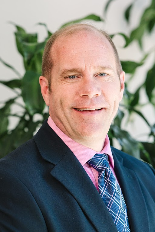 Portrait of a smiling man in a navy blazer, pink shirt, and blue patterned tie, posed indoors with green plants in the background.