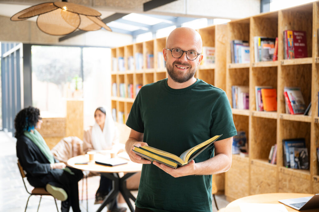 Studentsupport4 Antioch University Man holding an open book in a bright library or bookstore with people seated in the background